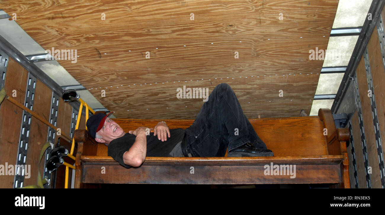 Mature man collapses on an old church pew that he is moving out of a moving  van. His face is covered with sweat and he is laying full length on the o