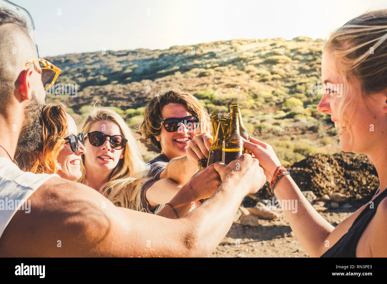 Group of young caucasian people boys and girls clinking together beer bottles in natural celebration day - enjoying outdoor and friendship lifestyle - Stock Photo