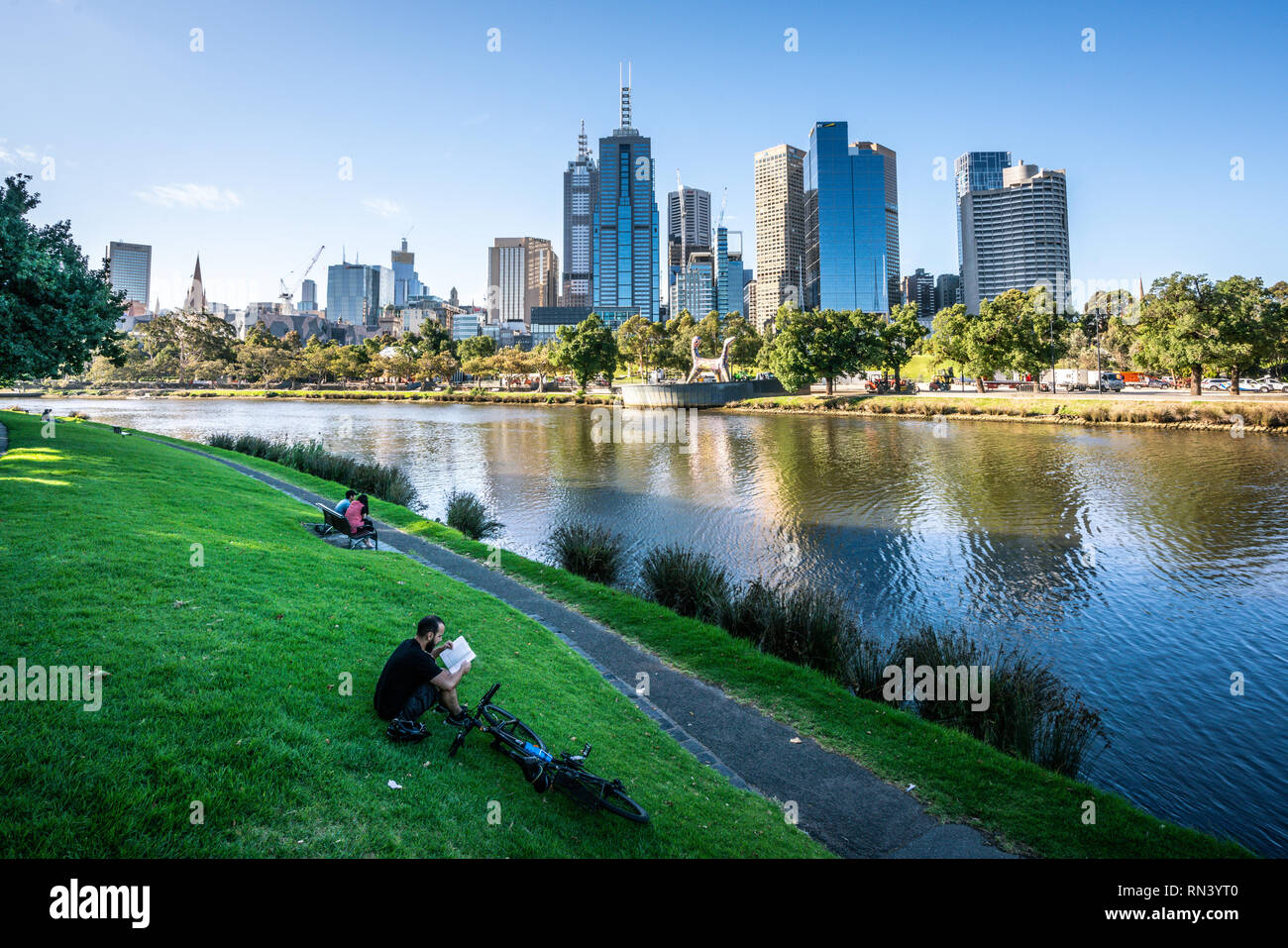 2nd January 2019, Melbourne Australia : Man reading a book on Yarra riverbank promenade grass in central Melbourne and Melbourne CBD skyline in Victor Stock Photo