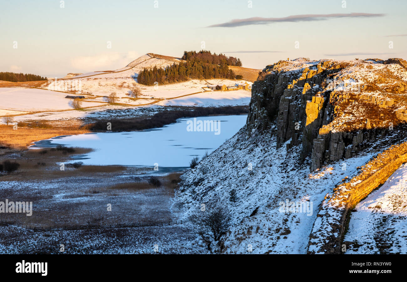 Winter snow lies on fields and crags beside the frozen lake of Crag Lough, while hikers walk along Hadrian's Wall path in Northumberland. Stock Photo
