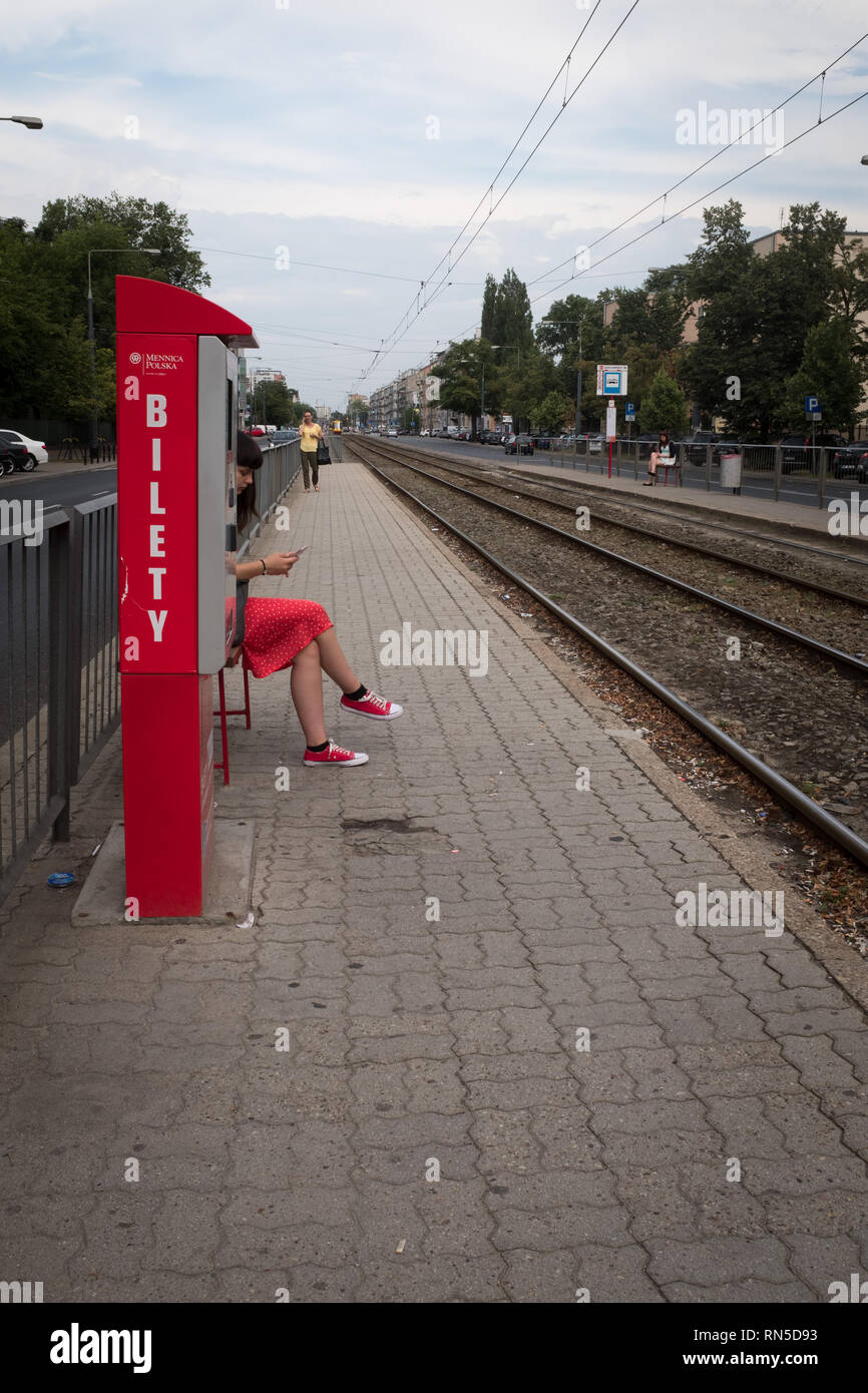 Woman in a red dress with red shoes sitting on a train platform hidden behind a red ticket machine Stock Photo