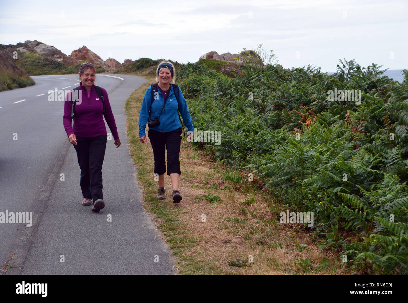 Two Women Hikers Walking, Talking & Laughing on Quiet Coastal Road near Cobo Bay on Guernsey, Channel Islands. UK. Stock Photo