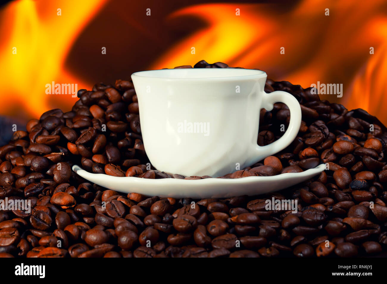 A white cup on top of a pile of roasted coffee beans with fire in the background. Stock Photo