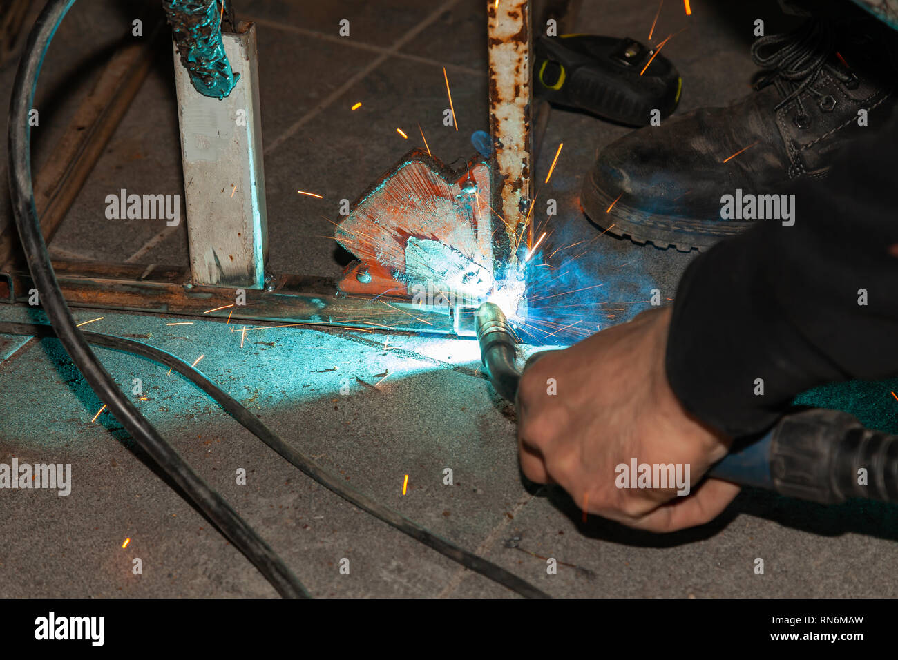 An experienced person performs work with a welding machine, fixing metal parts, removing blue smoke and yellow sparks and lightning in the industrial  Stock Photo
