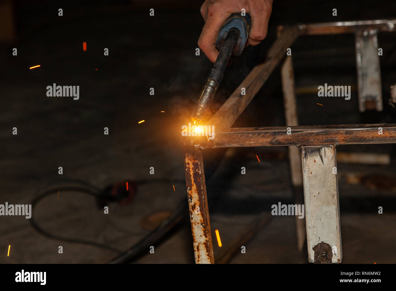 An experienced person performs work with a welding machine, fixing metal parts, removing blue smoke and yellow sparks and lightning in the industrial  Stock Photo
