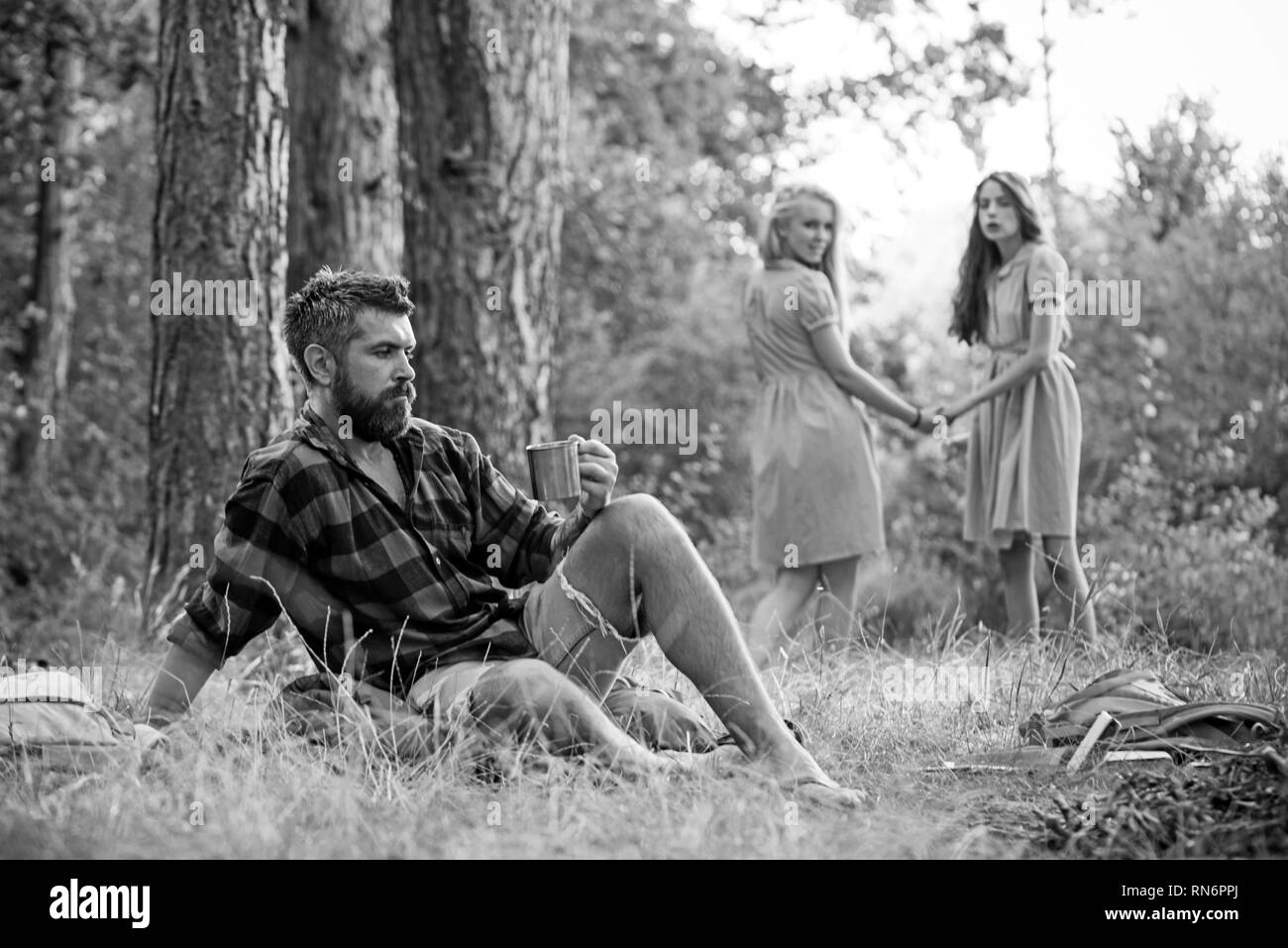 Friends camping in forest. Two girls holding hands look back at man sitting on grass. Side view bearded guy sitting on green meadow Stock Photo