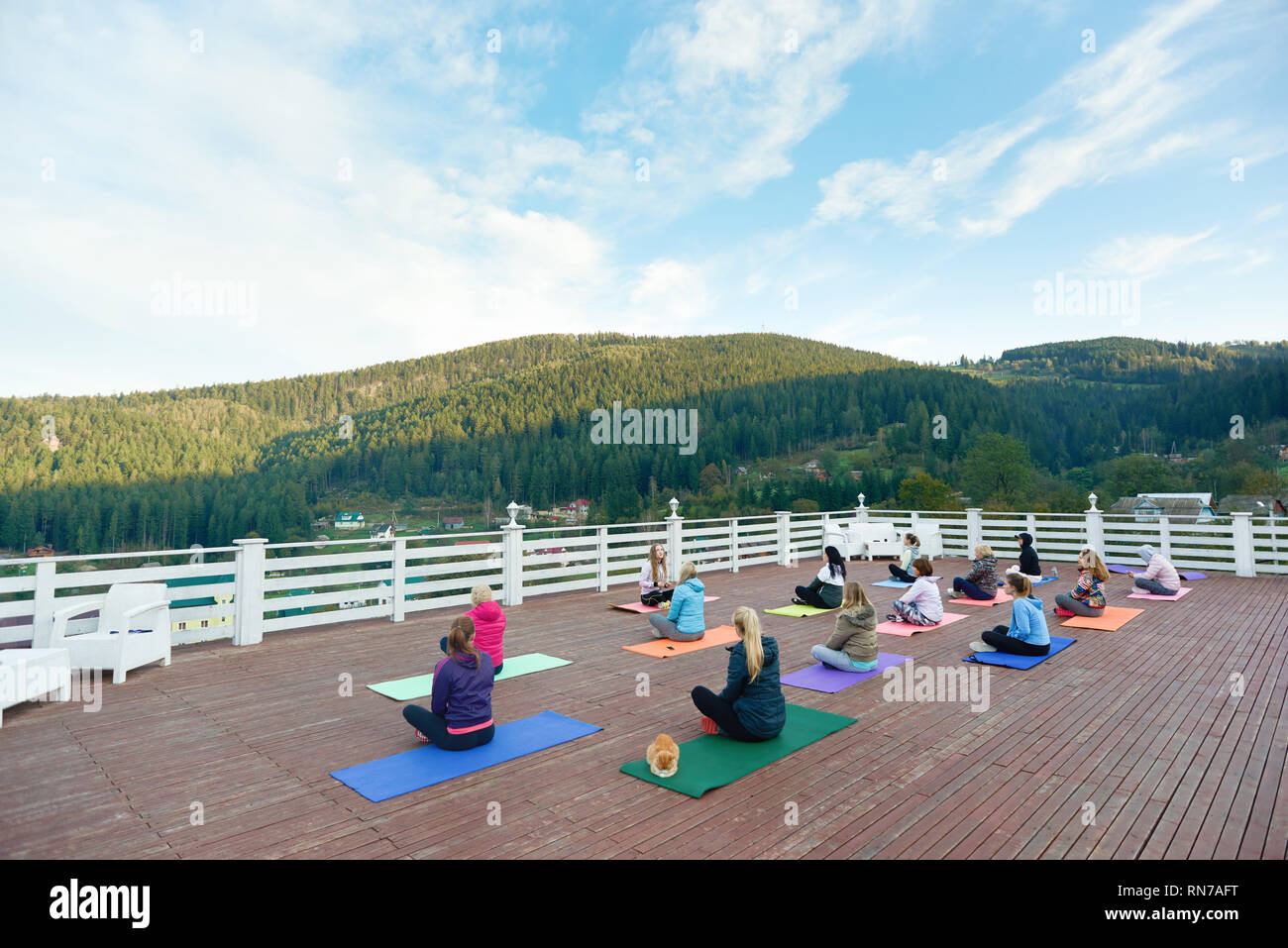 Group of people doing yoga together with coach on fresh air. People sitting in lotus pose observing amazing view of mountains. Women practicing on yoga mats, wearing in sportswear. Stock Photo