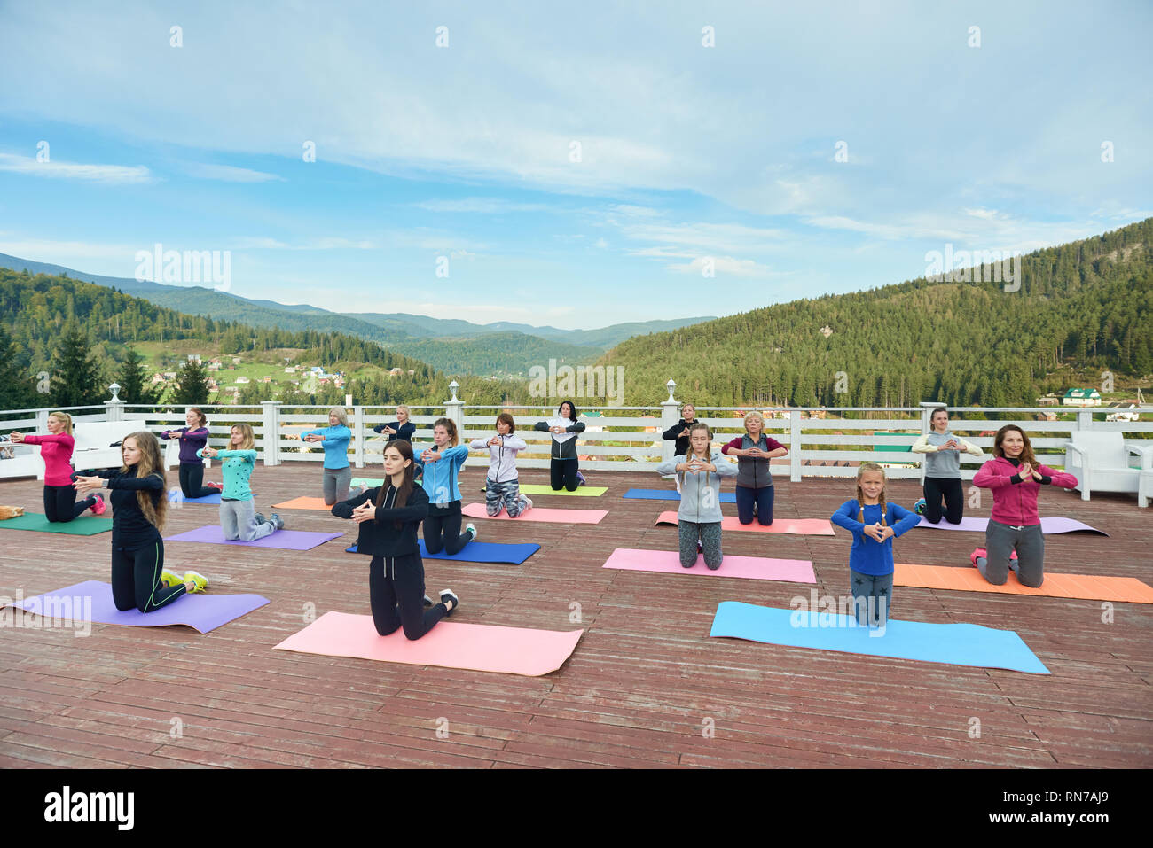 Female yoga group practicing yoga asana outdoors in mountains under blue sky. Women standing on knees and putting hands. People using yoga mats for doing exercises. Stock Photo