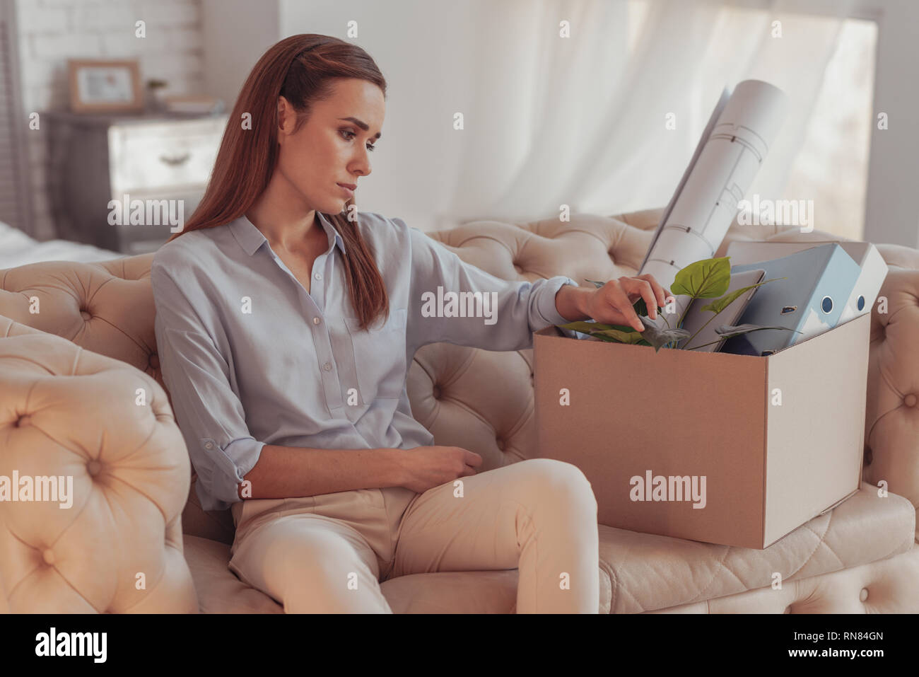 Again unemployed. Young dispirited woman sitting on sofa and being lost in thoughts while touching a box with her office property Stock Photo