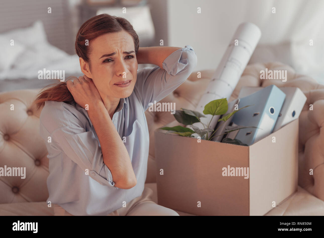 Job problems. Portrait of upset young woman looking away and touching her neck while crying on the sofa with the stuff from her former work Stock Photo