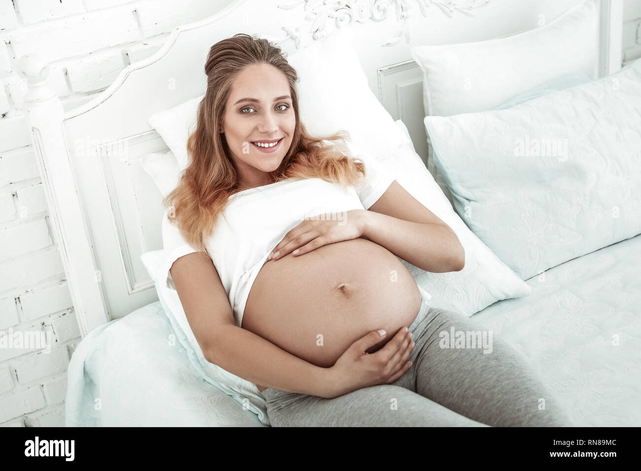 Pleasant woman with wide smile wrapping pregnant belly Stock Photo