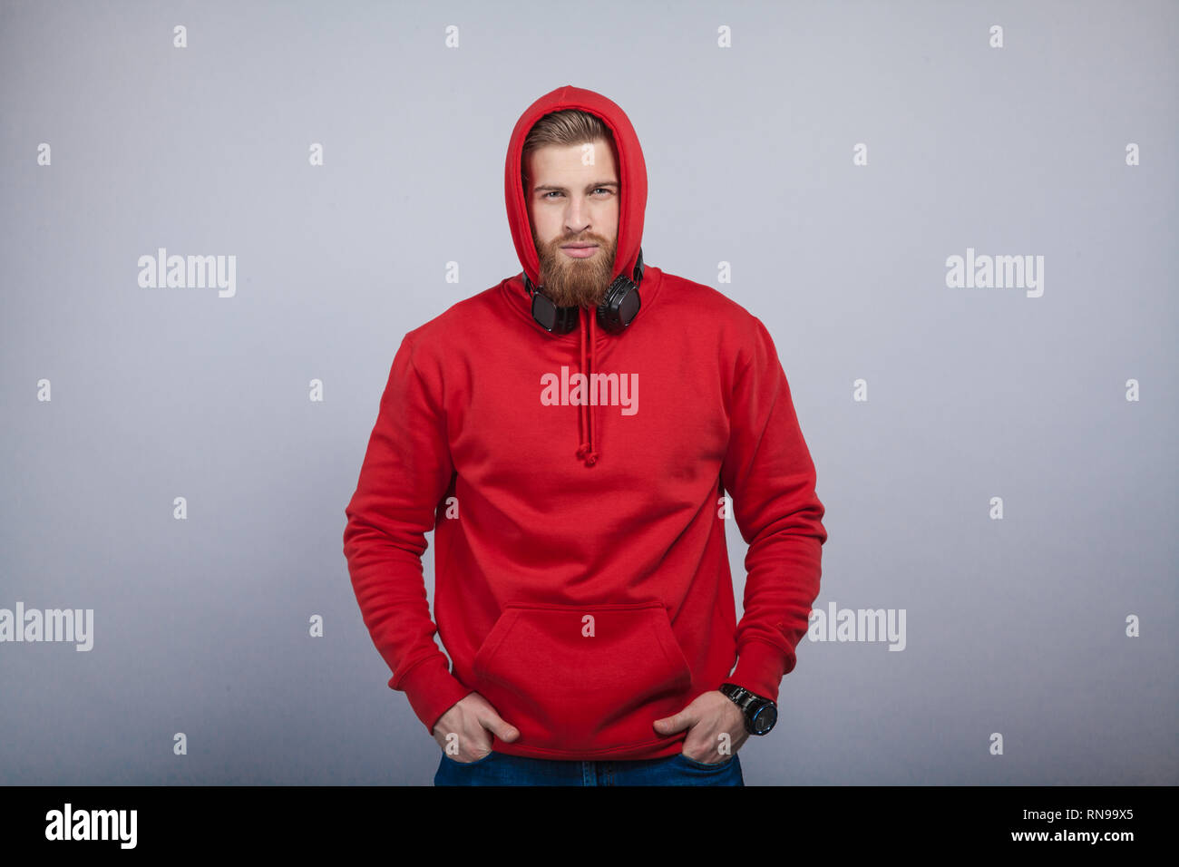 young handsome man with the beard wearing red sweatshirt with hood on and headphones on his neck standing at the grey background looking to the camera Stock Photo