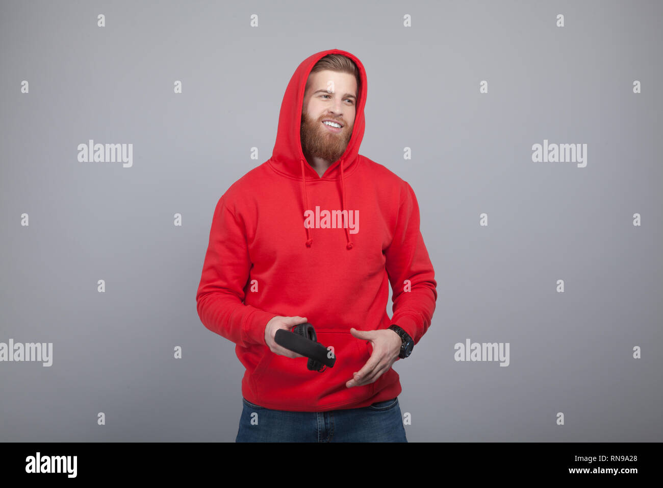 young handsome smiling bearded man in red hoodie on holding headphones in his hands standing at the grey background and looking to the camera. Stock Photo