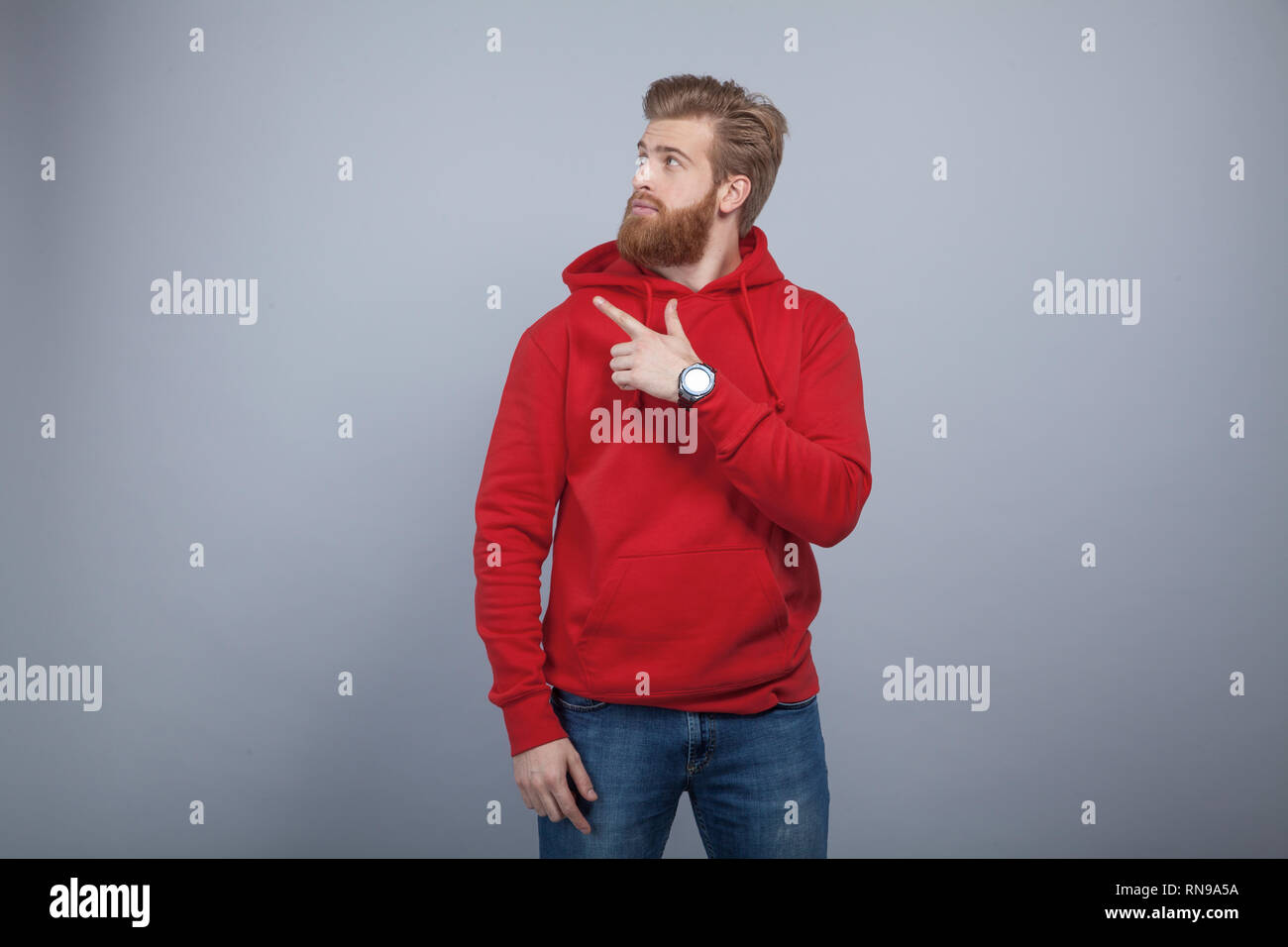 young handsome bearded man wearing red sweatshirt standing on the grey background and pointing his finger on the left side and looking there with the  Stock Photo