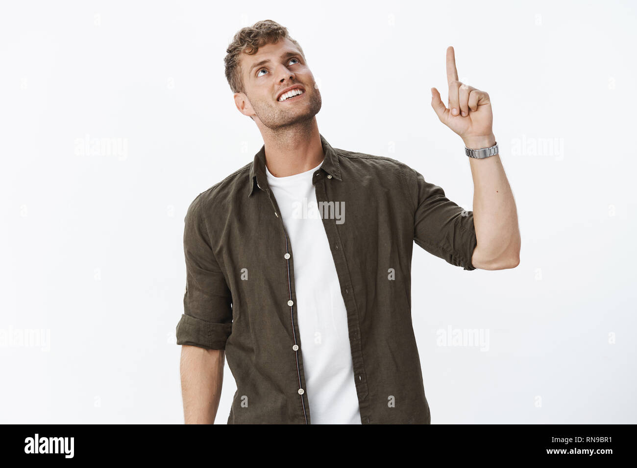 Delighted happy and pleased handsome blond guy with blue eyes and bristle wearing watch, shirt over t-shirt looking and pointing up with joyful smile Stock Photo