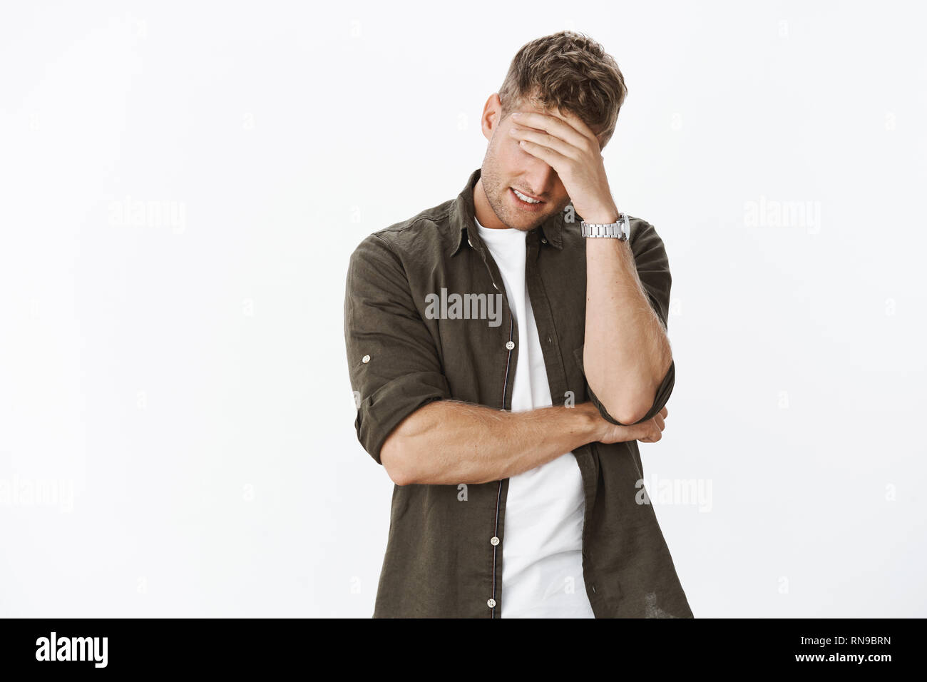 Embarrassed and awkward handsome ex-boyfriend trying hide behind hand as making facepalm gesture so girl would not recognize him, smiling from stress Stock Photo