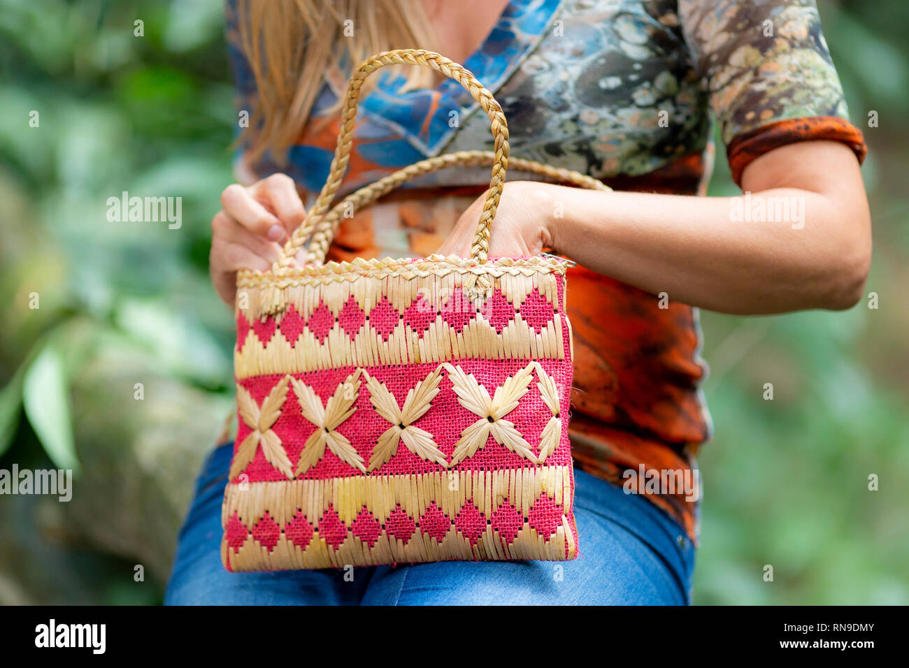 Closeup of the hands of a woman in jeans and colourful top reaching in her handbag that rests on her knees in a natural surrounding in the background Stock Photo
