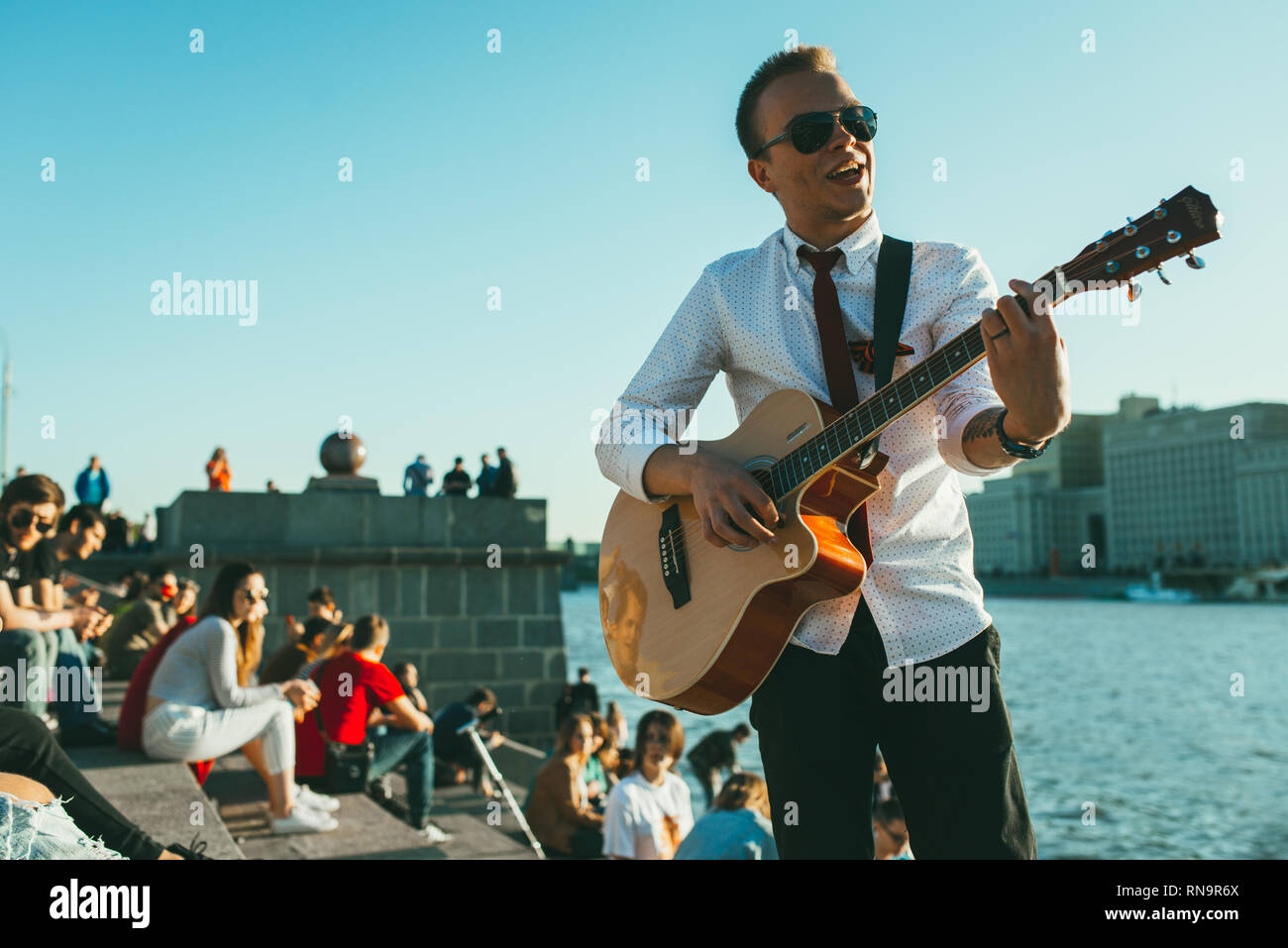 Moscow, Russia - May 9, 2018: young man with sunglasses, tie and St. George Ribbon plays guitar against the riverside. Crowd of people sitting and Stock Photo