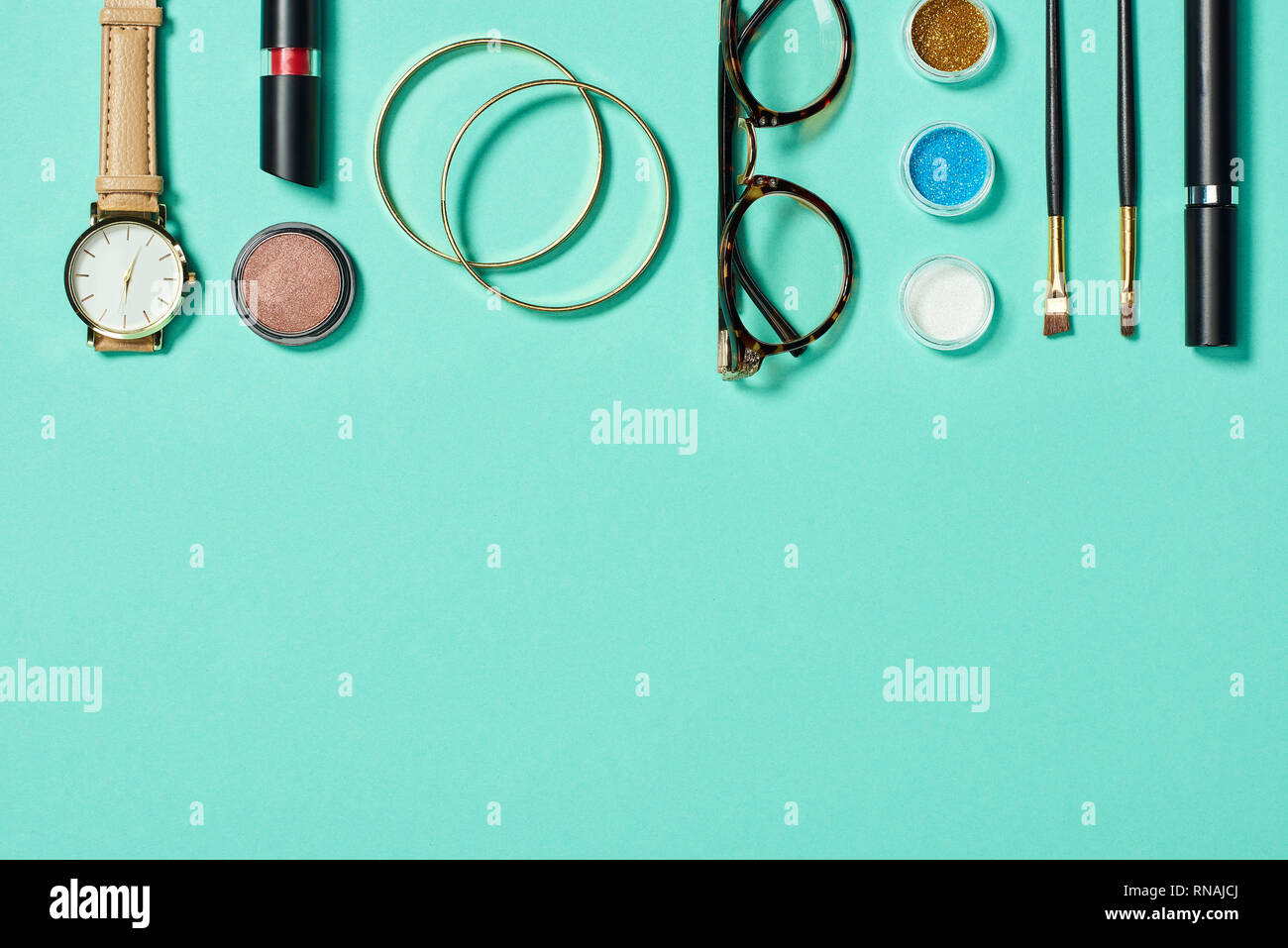 Top view of watch, lipstick, bracelets, glasses, eyeshadow, blush, cosmetic brushes and mascara on turquoise background Stock Photo