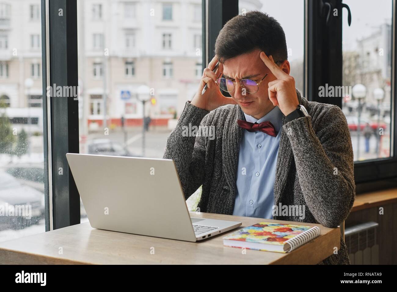 Young tired, ill, overworked man in formal wear sitting in front of computer and touching his forehead Stock Photo