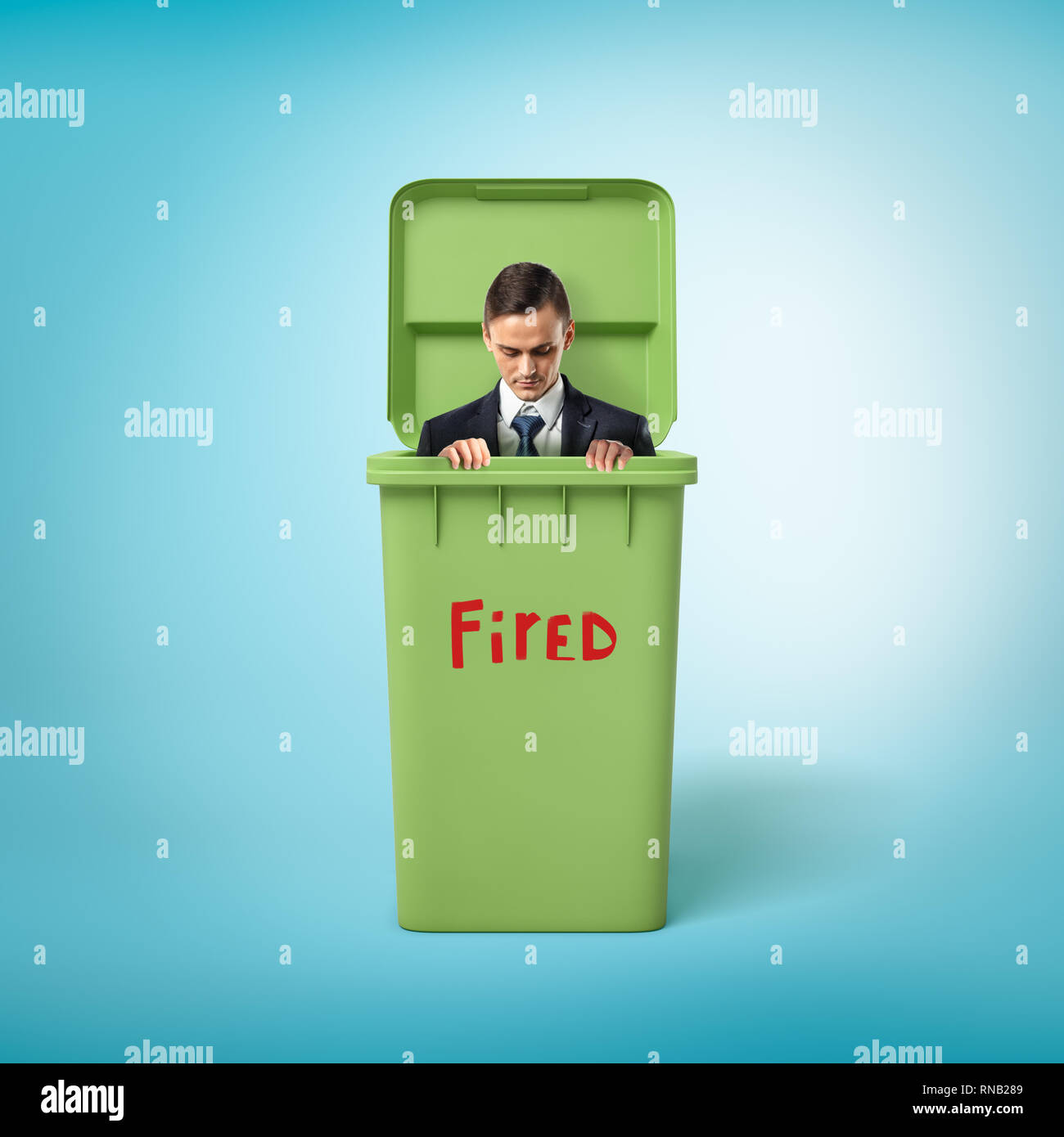 Young businessman in green trash bin with the red 'FIRED' sign on blue background Stock Photo