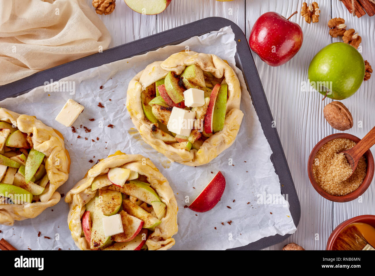 open apple pies, galettes with apple slices stuffing topped with butter cubes, prepared to bake on a baking tray. apple, cinnamon sticks and kitchen c Stock Photo