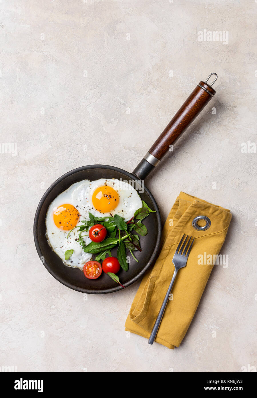 Black pan with fried eggs, fresh herbs and cherry tomatoes, fork and napkin at white background. Concept of healthy food. Top view, free space Stock Photo