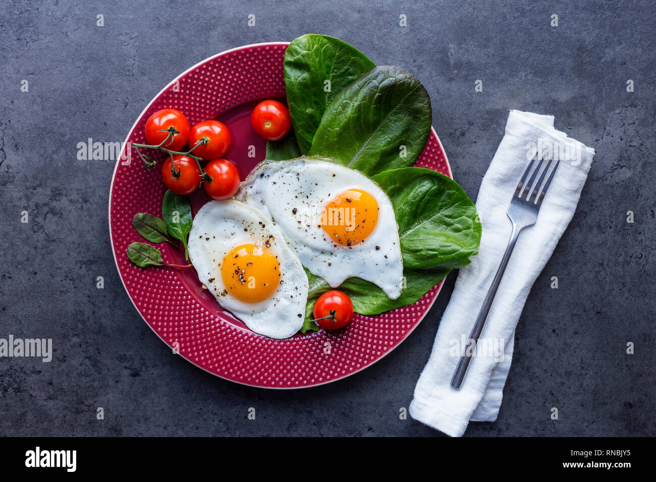 Serving plate with fried eggs, salad and tomatoes, fork and napkin at black background. View from above, cover for magazine Stock Photo