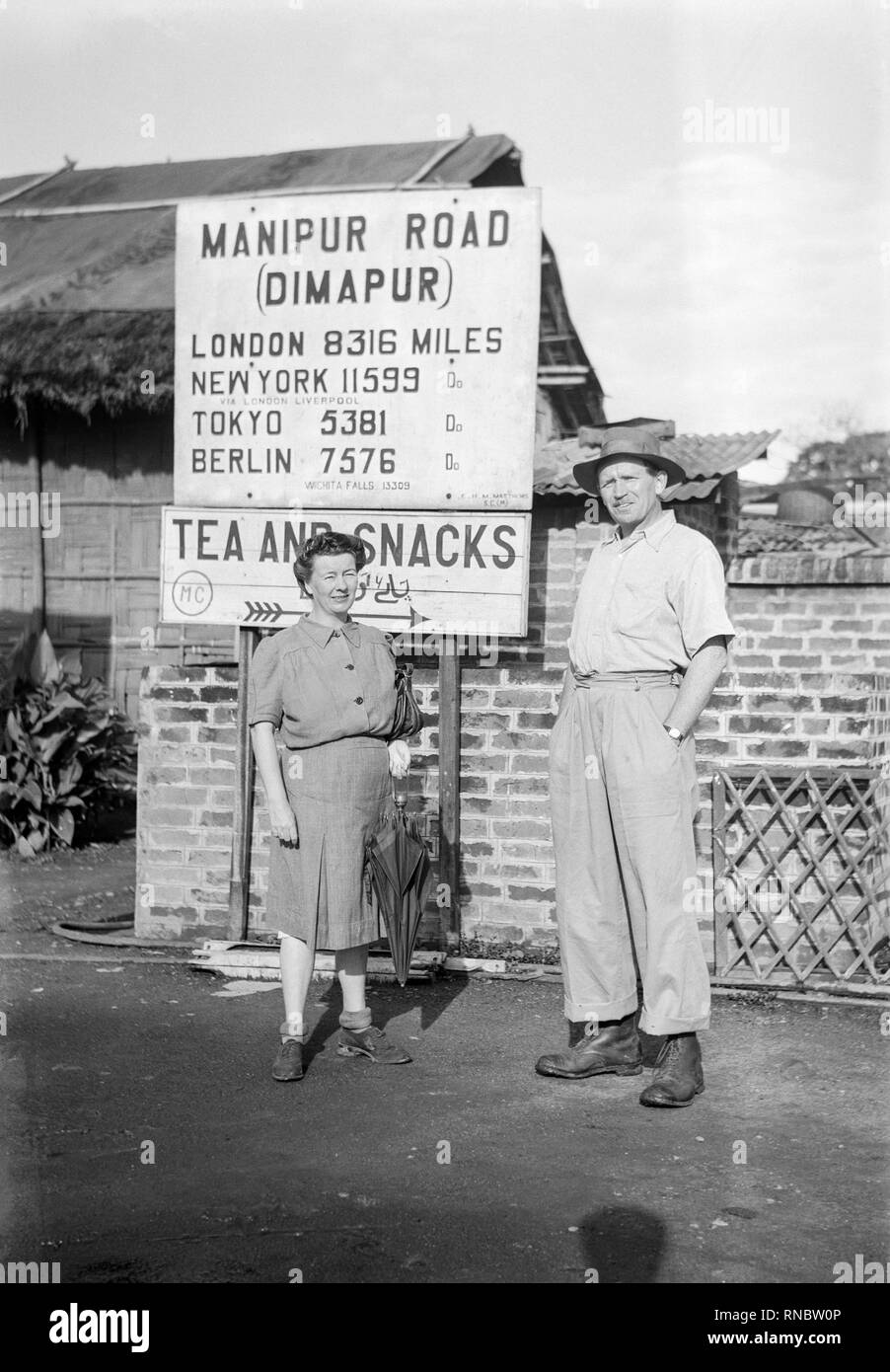A middle age white European man and woman posing in front of a sign for the Manipur Road in Dimapur, India. Manipur Road in Dimapur (State of Nāgāland) is a city located in India about 1,033 mi (or 1,663 km) east of New Delhi, the country's capital town. Vintage mid twentieth century black and white photograph. Sign gives distances to various cities around the world. Stock Photo