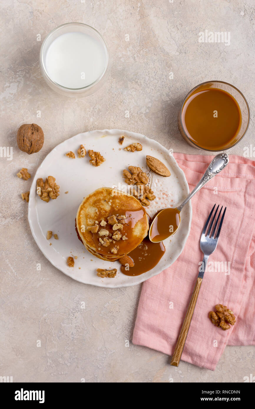 Stack of baked pancakes with boiled condensed milk and walnuts, almond on white plate with fork, knife, pink napkin, jar of caramel and milk in glass  Stock Photo
