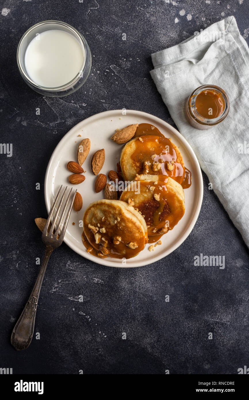 American pancakes with caramel and nuts at white plate, fork, napkin, jar with caramel and glass of milk near at black background. Concept of healthy  Stock Photo