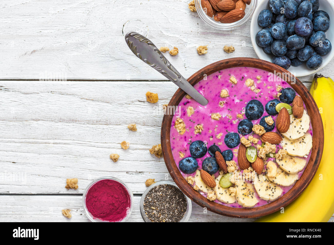 Acai smoothie bowl topped with banana, chia and pumpkin seed, blueberry, almonds and granola with spoon on white wooden background. healthy vegan brea Stock Photo