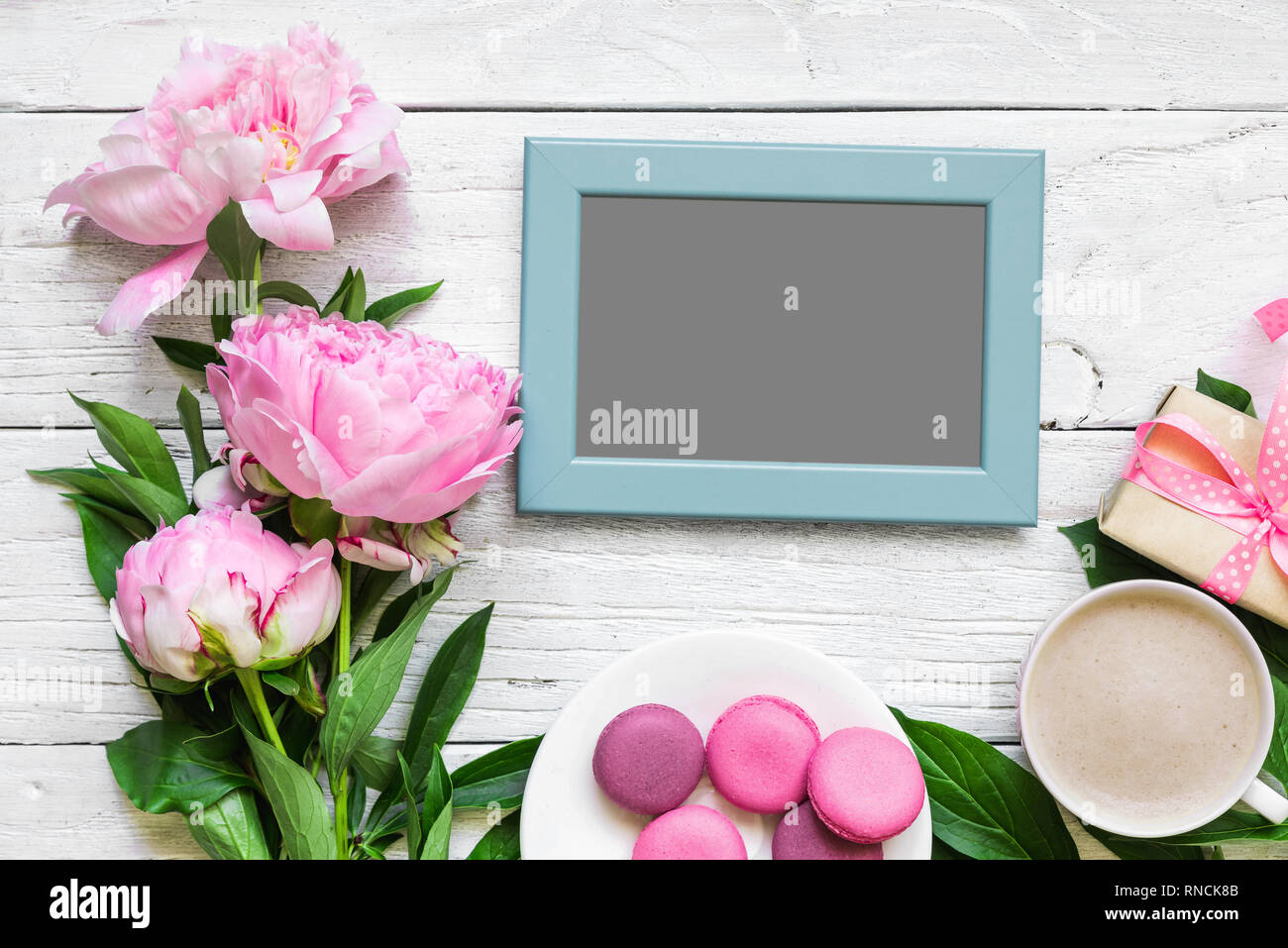 blank photo frame and peony flowers with cappuccino cup, macaroons and gift box on white wooden table. mock up. Flat lay. wedding or womens day concep Stock Photo