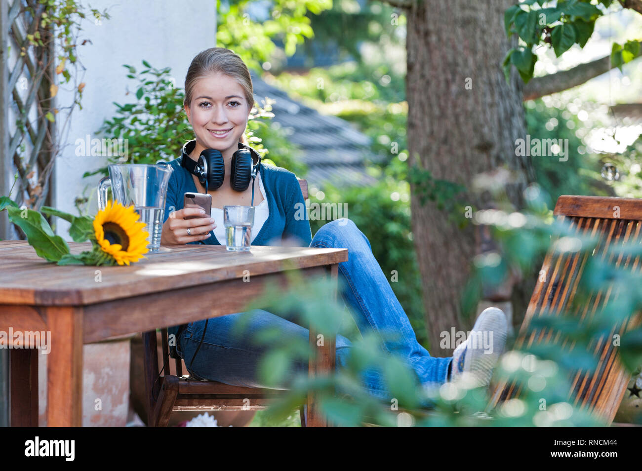 Junge Frau mit langen blonden Haaren sitzt im Garten an einem hoelzernen Tisch mit einem Handy in der Hand und Kopfhoerern  [(c) Dirk A. Friedrich Stock Photo