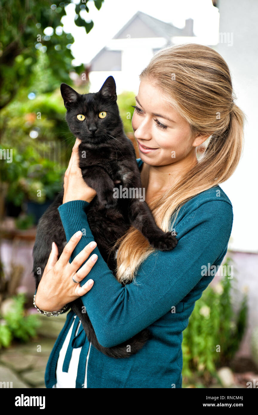 Junge Frau mit langen, blonden Haare steht im Garten und haelt eine schwarze Katze im Arm [(c) Dirk A. Friedrich Stock Photo