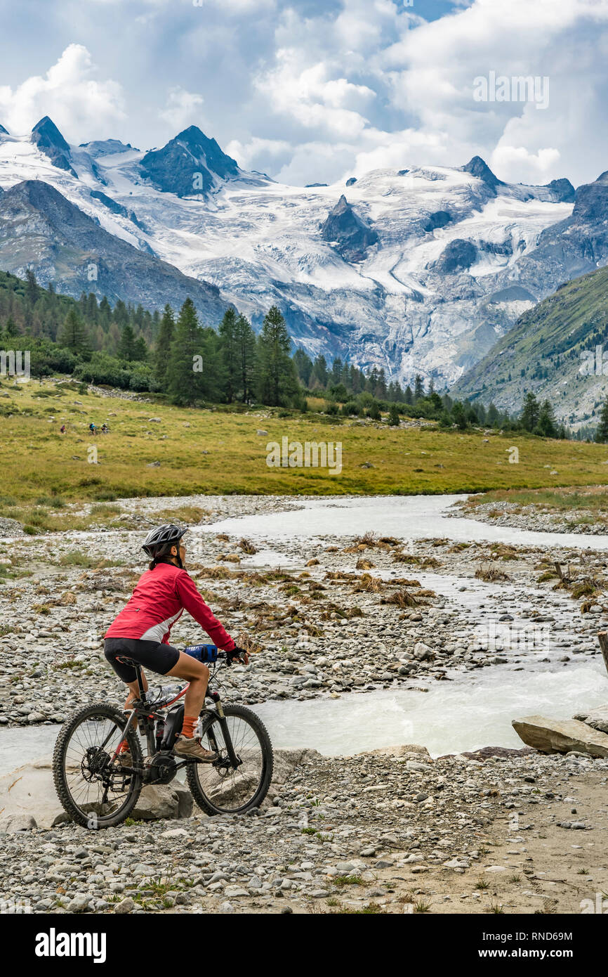 active senior woman, riding her e-mountain bike in the Roseg valley below the glaciers and summits of the Sella Group and Piz Roseg Stock Photo