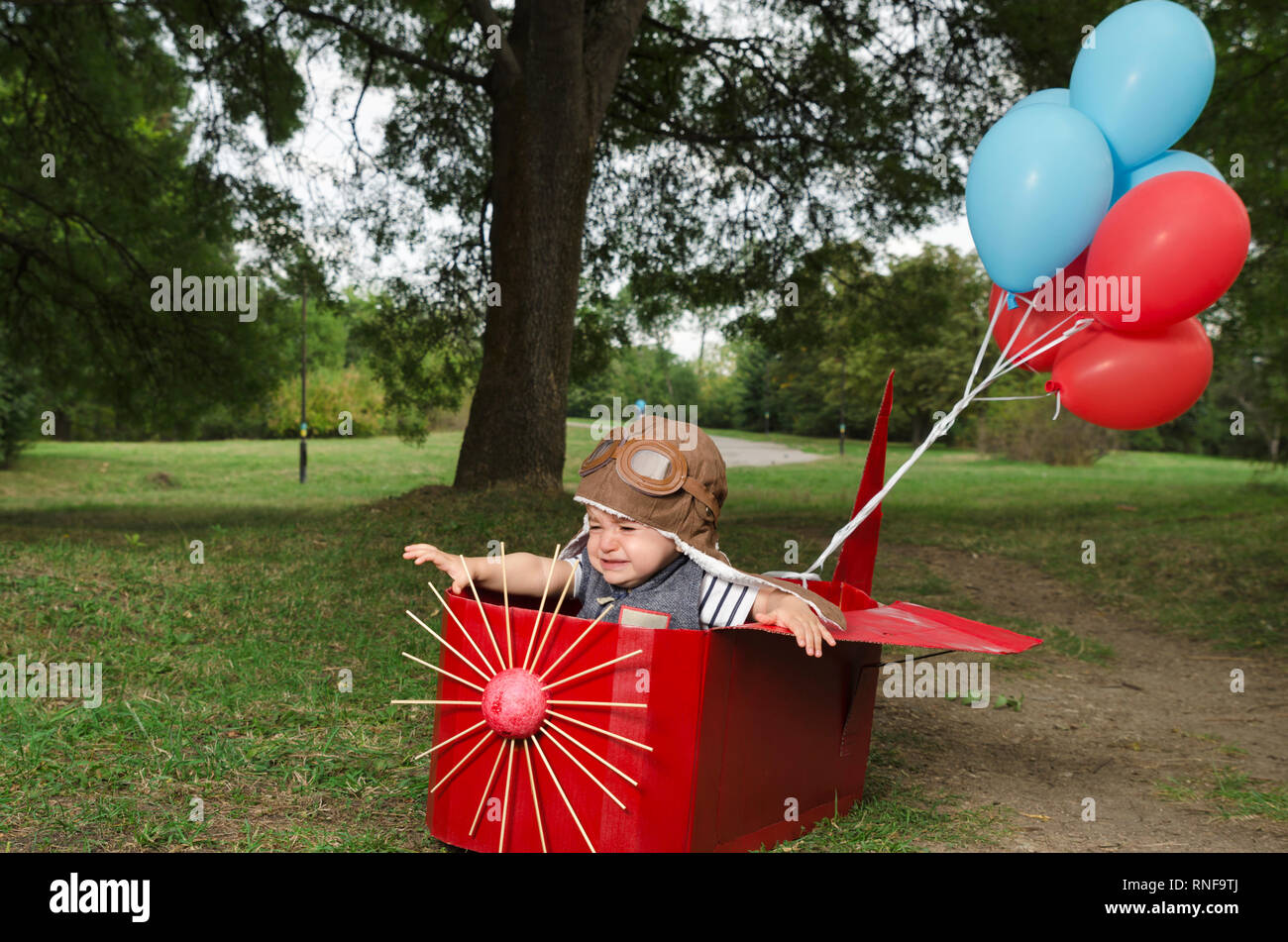 Cute toddler with aviator hat crying in a red paper handmade airplane with balloons. Selective focus Stock Photo