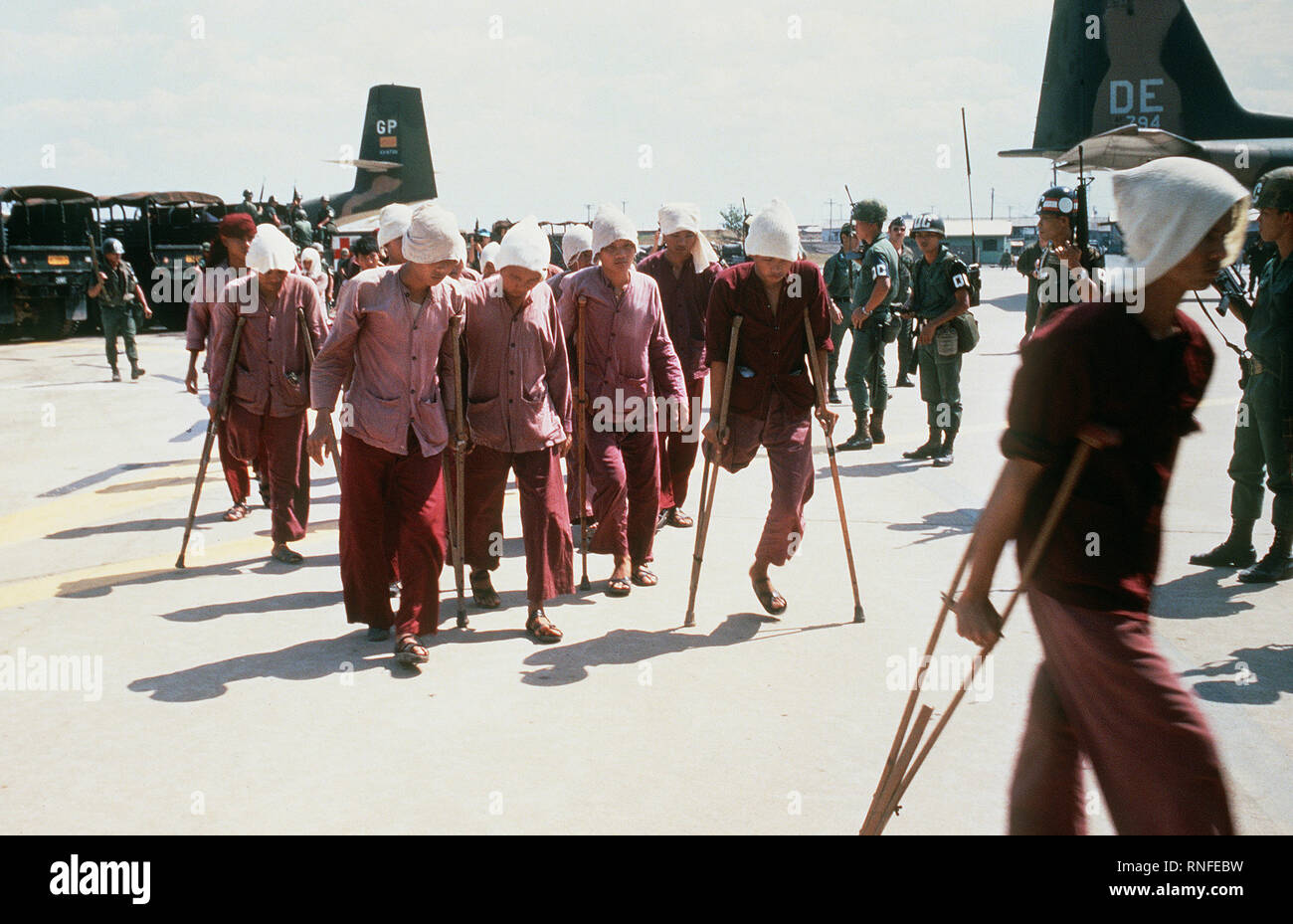 Viet Cong POWs, some on crutches, under the watchful eyes of South Vietnamese military police walk to the waiting C-123 transport aircraft.  The POWS will be airlifted to Loc Ninh, South Vietnam for the prisoner exchange between the United States/South Vietnam and North Vietnam/Viet Cong militaries. Stock Photo