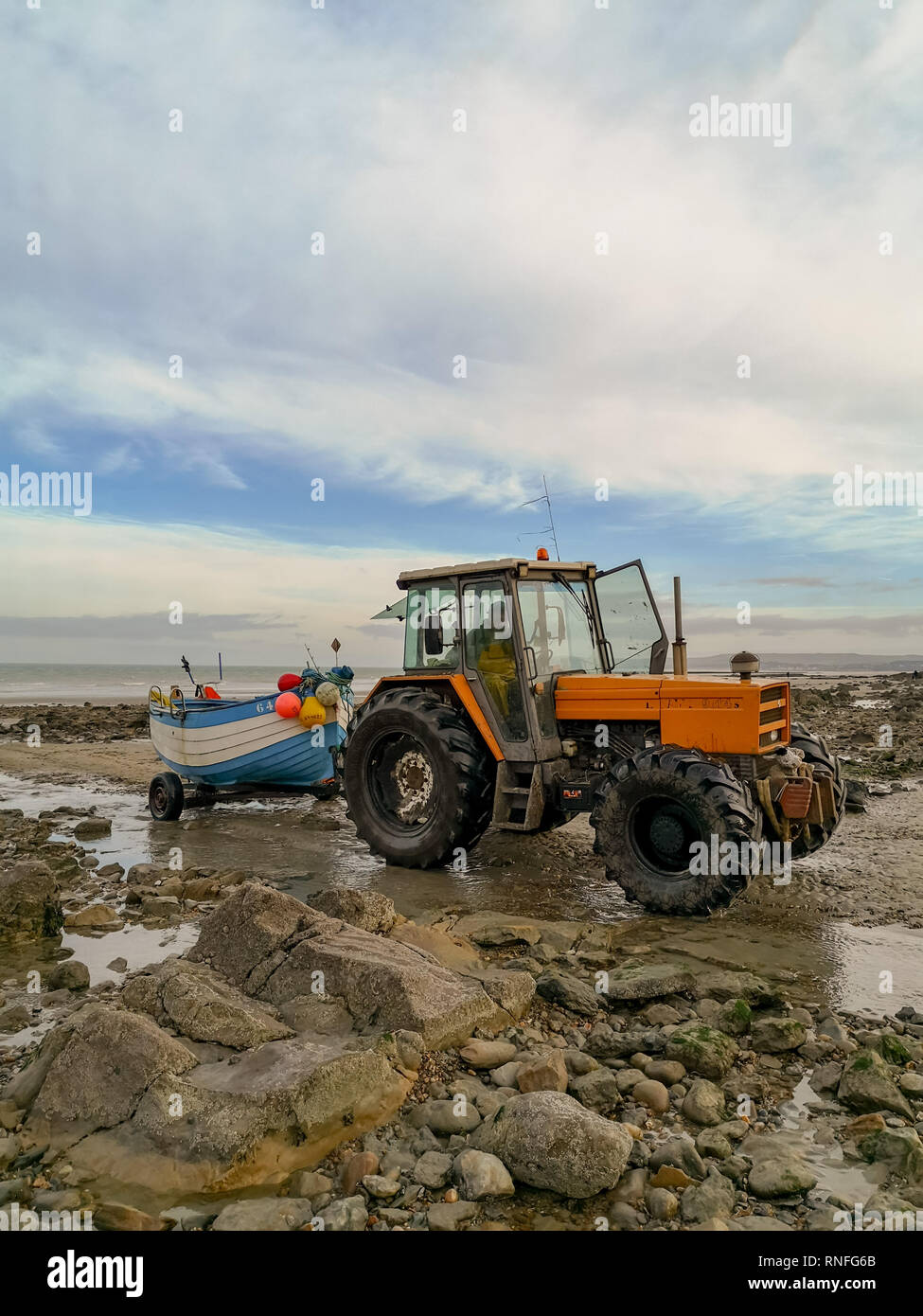 December 2018 - Wissant, France: Orange tractor towing a wooden white and blue fishing boat on a rocky beach during low tide Stock Photo