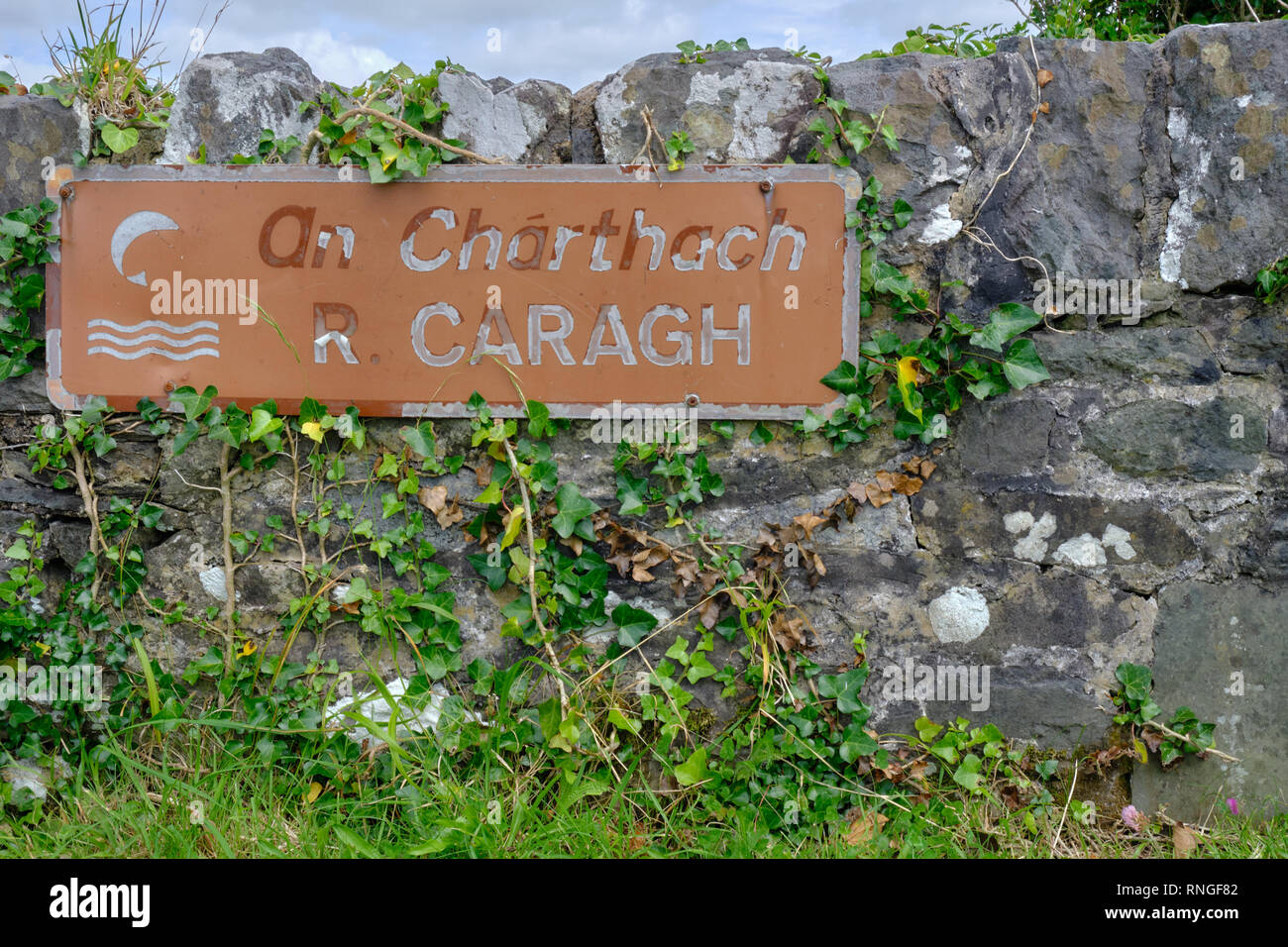 Sign on low road wall for River Caragh, both in English and in Gaelic.  Vine growing on wall and sign Stock Photo