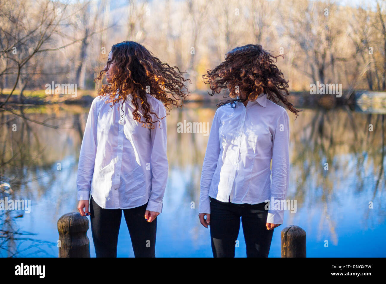 Young twin sisters in same clothes shaking hair at lake on autumn day ...