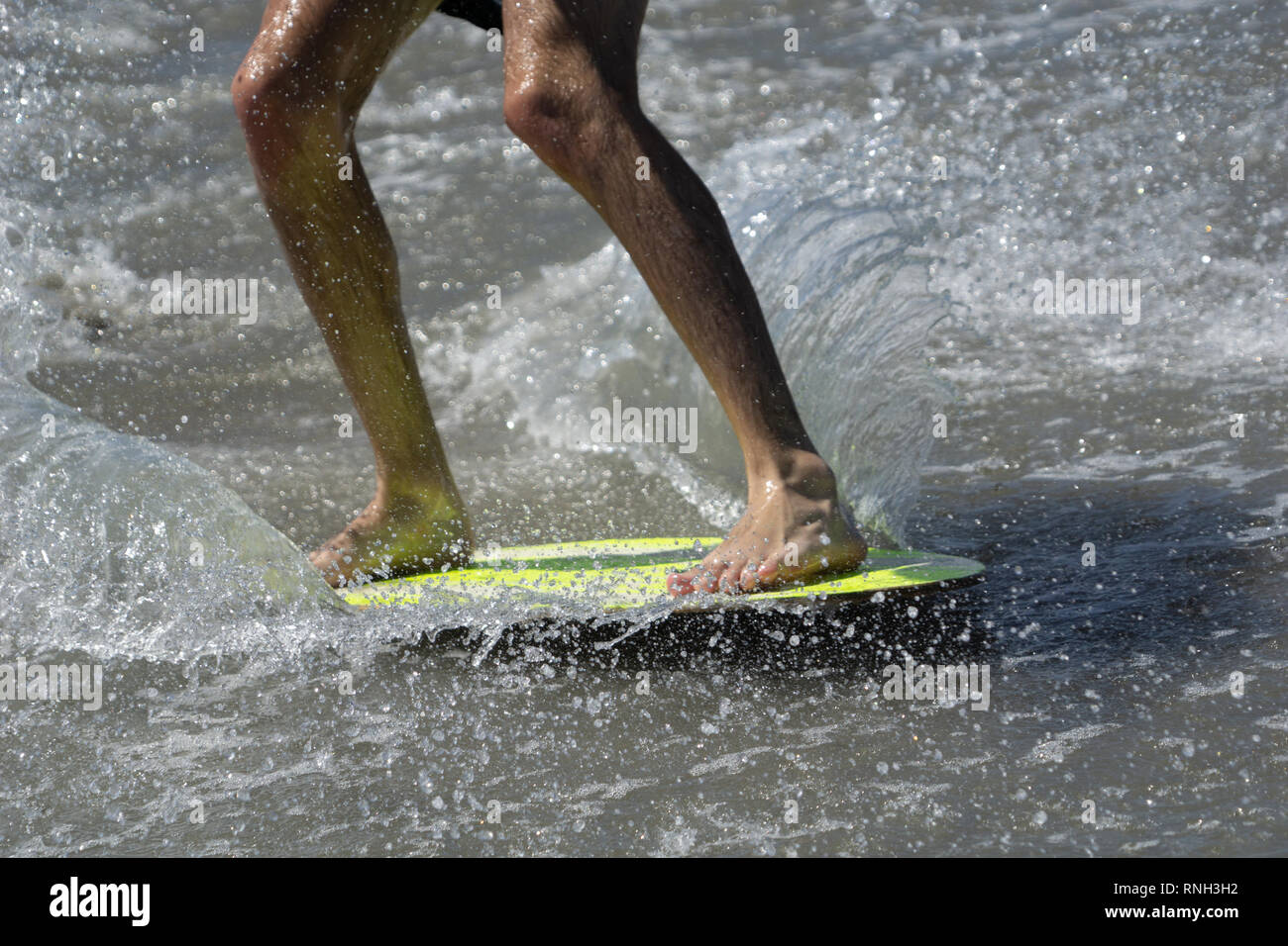Skim-boarding mans legs close-up on yellow board in shallow water. Stock Photo