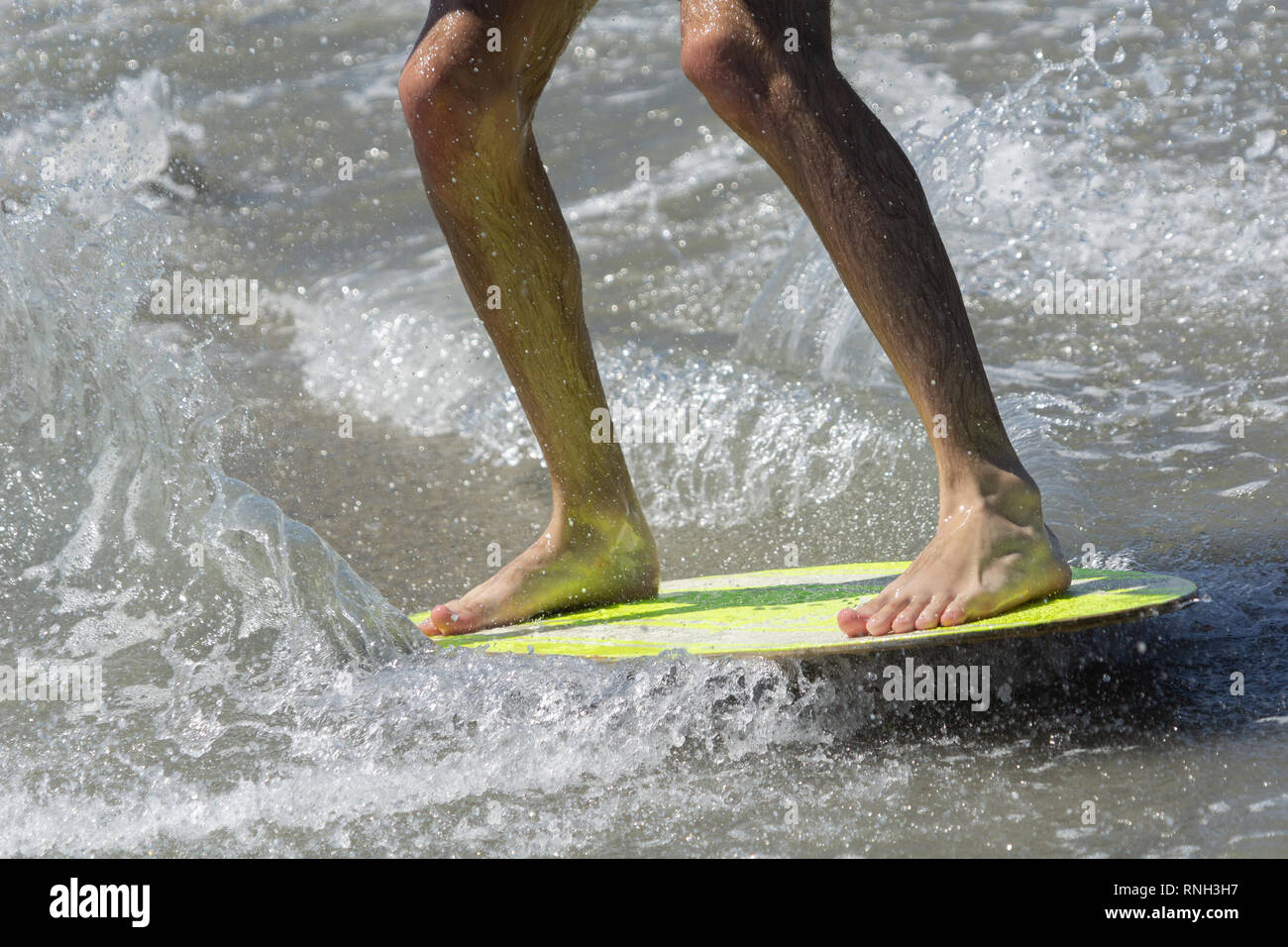 Skim-boarding mans legs close-up on yellow board in shallow water. Stock Photo