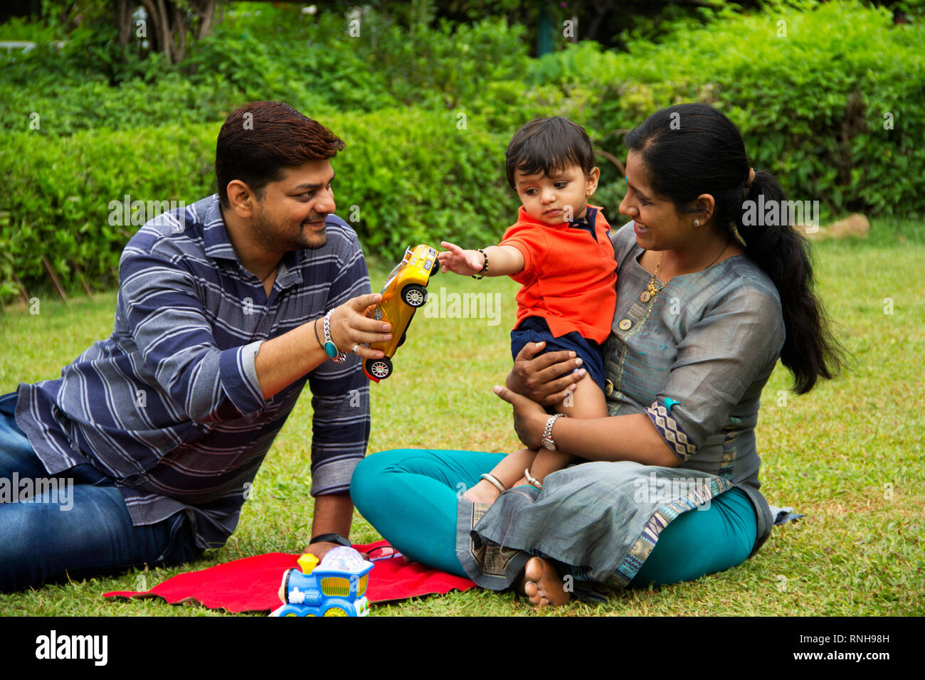 Happy Indian family, father giving toy car to his toddler son, sitting with his mother, inside garden, Pune, Maharashtra Stock Photo