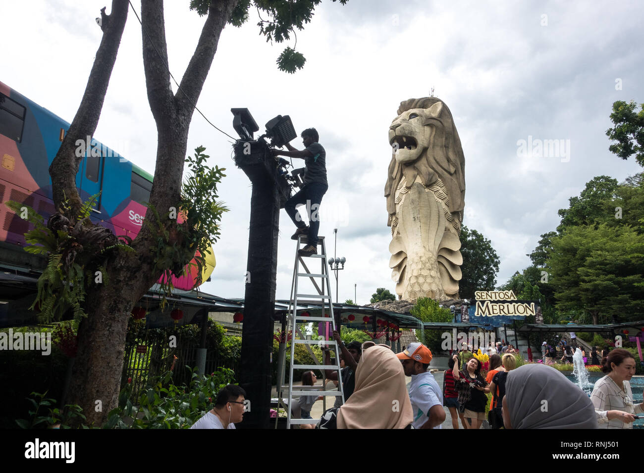 Singapore, Singapore - January 22, 2017: A worker repairs lighting on a ladder, with iconic merlion statue and tourists in the distance, Sentosa Islan Stock Photo