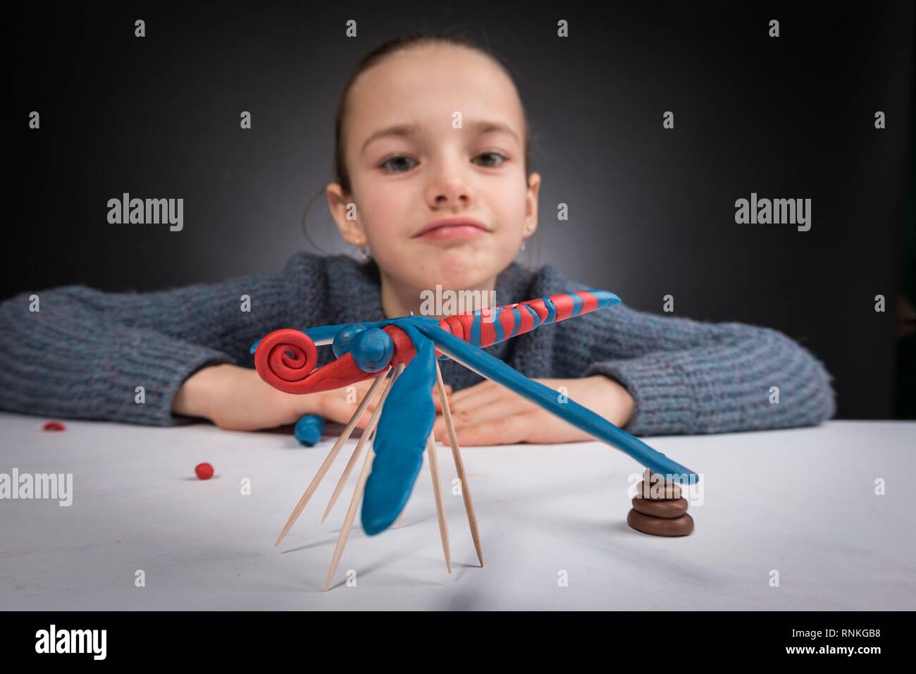 wide-angle portrait of a teenage girl 9-12 years old in defocused form, in the foreground on a white table stands a dragonfly of blue-red pastelina, w Stock Photo