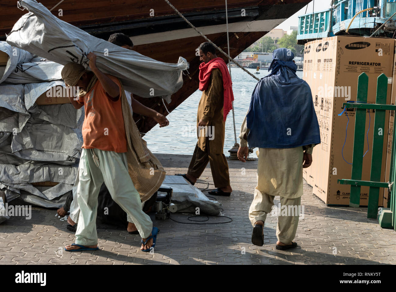 Asian  dock workers carrying large goods be loaded onto a moored Dhow ( Arab boat) on the quayside of the Dubai Creek in Dubai, United Arab Emirates,  Stock Photo