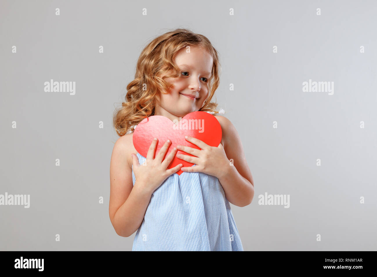 Portrait of an adorable little child girl cuddles a red paper heart. Gray background, studio Stock Photo