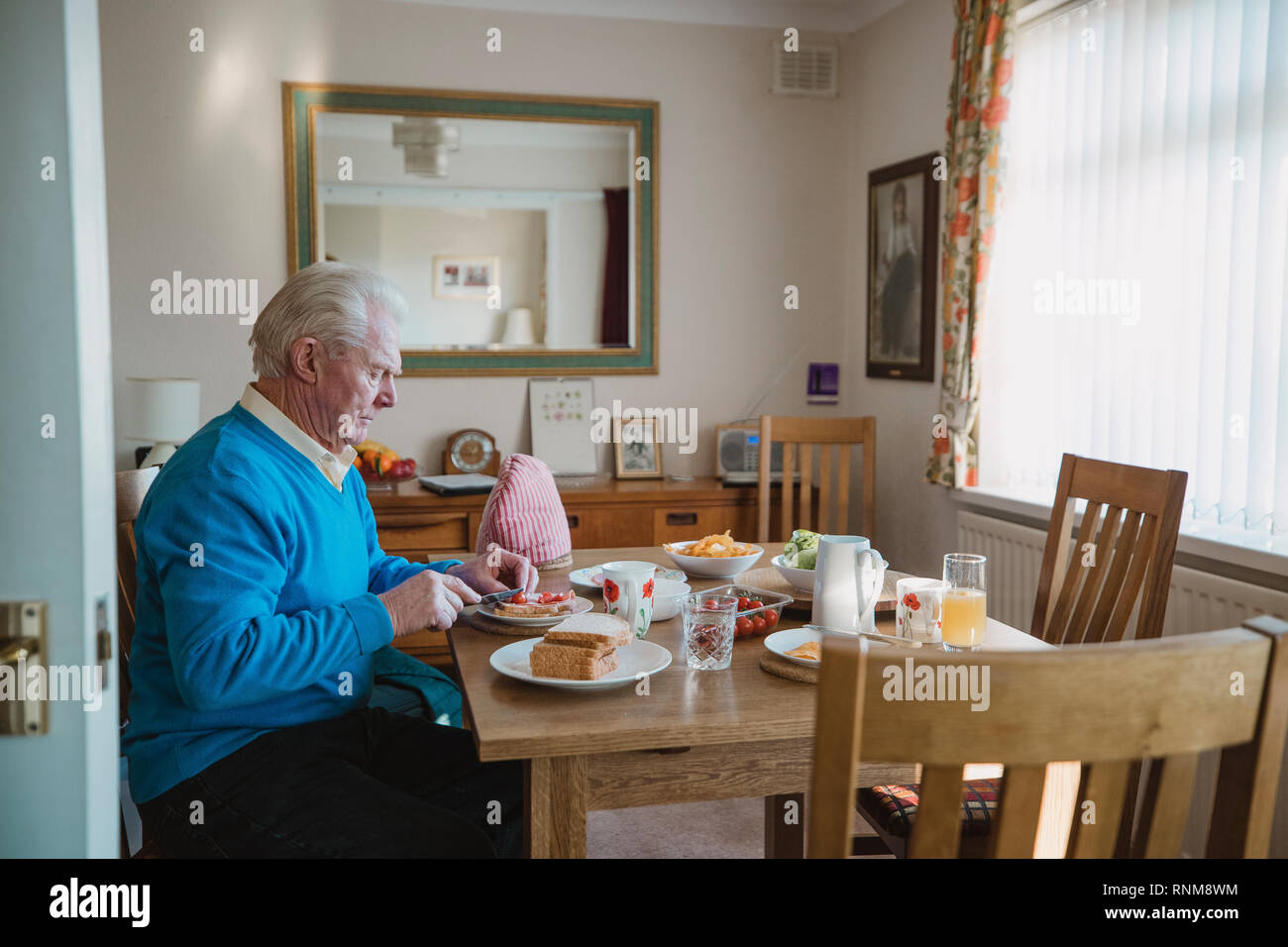 One senior man is sitting at the dining table in his home having lunch. He is making a ham salad sandwich. Stock Photo