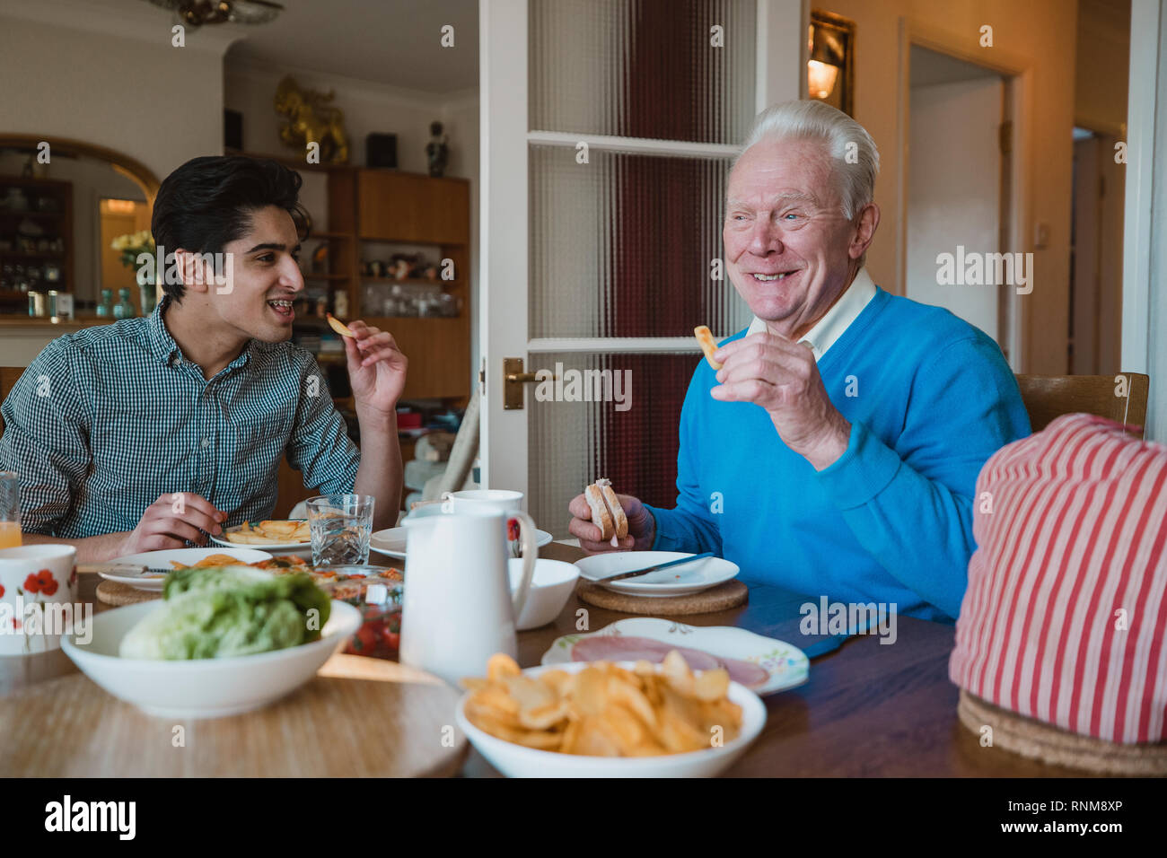 Senior man is having lunch at home with his grandson. They are eating sandwiches and potato chips. Stock Photo